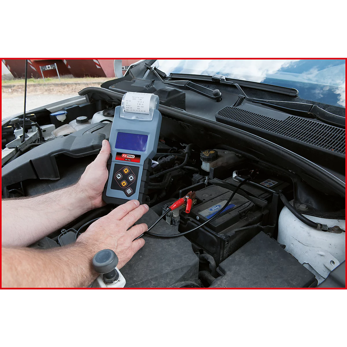 Person testet Autobatterie mit einem Messgerät. Der Motorraum ist geöffnet, Messklemmen sind an der Batterie befestigt.